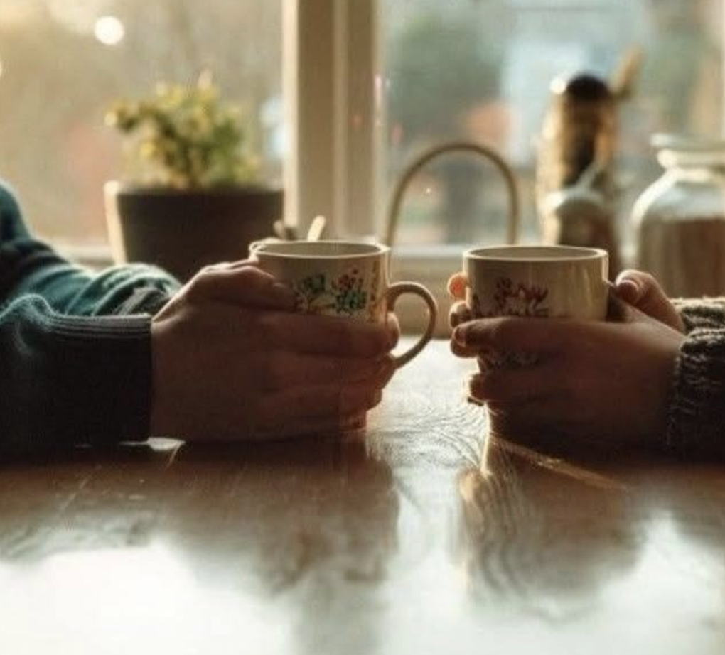 Two friends across a kitchen table holding floral coffee mugs in warm window light